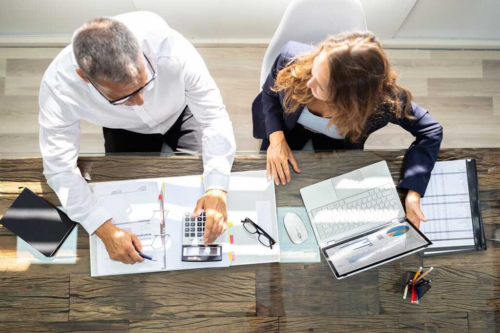Two professionals seated at a desk reviewing financial documents with a calculator and laptop in an office setting.