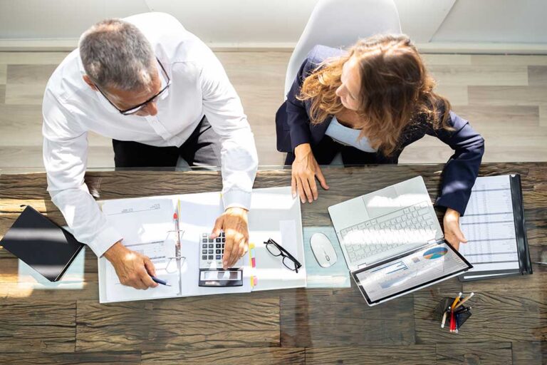 Two professionals seated at a desk reviewing financial documents with a calculator and laptop in an office setting.